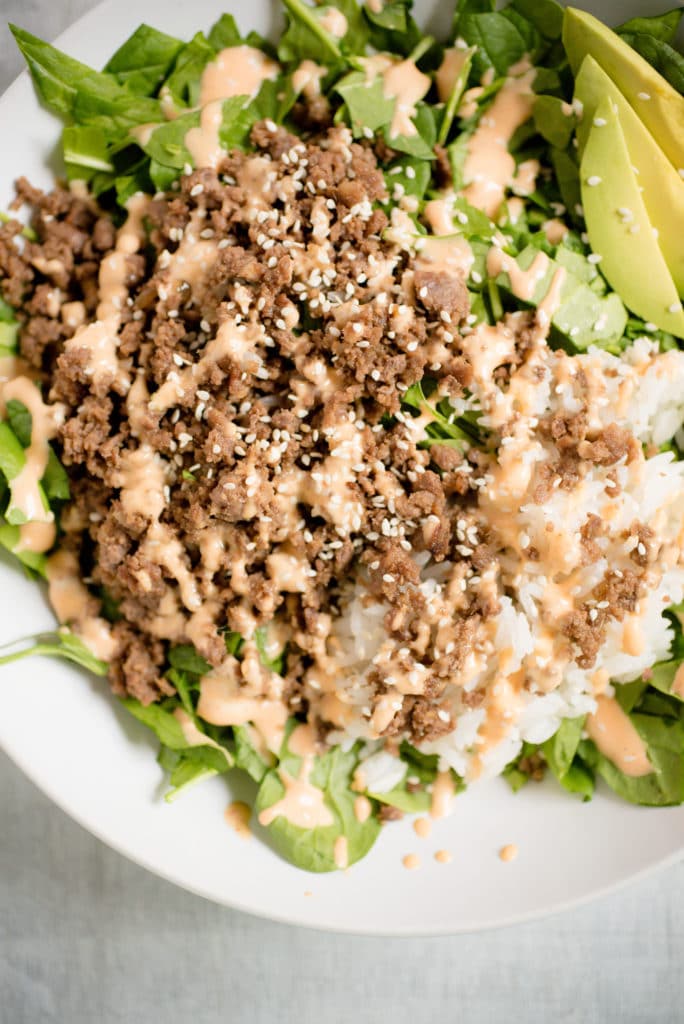 a bed of greens with avocado, korean beef, rice, spicy mayo, and white sesame seeds