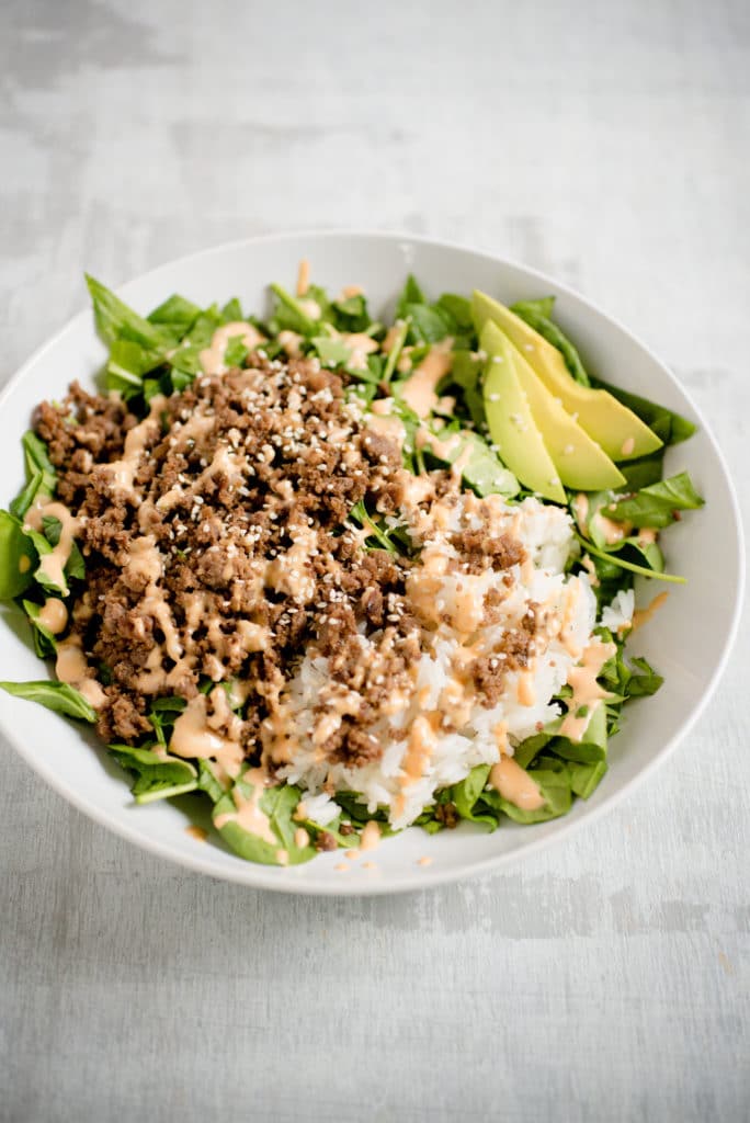 korean beef in a white bowl on top of lettuce greens with sliced avocado and rice. it is topped with spicy mayo and white sesame seeds