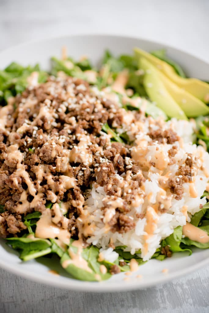 detailed view of greens topped with korean beef, rice, white sesame seeds, and spicy mayo with slices of avocado seen in the background