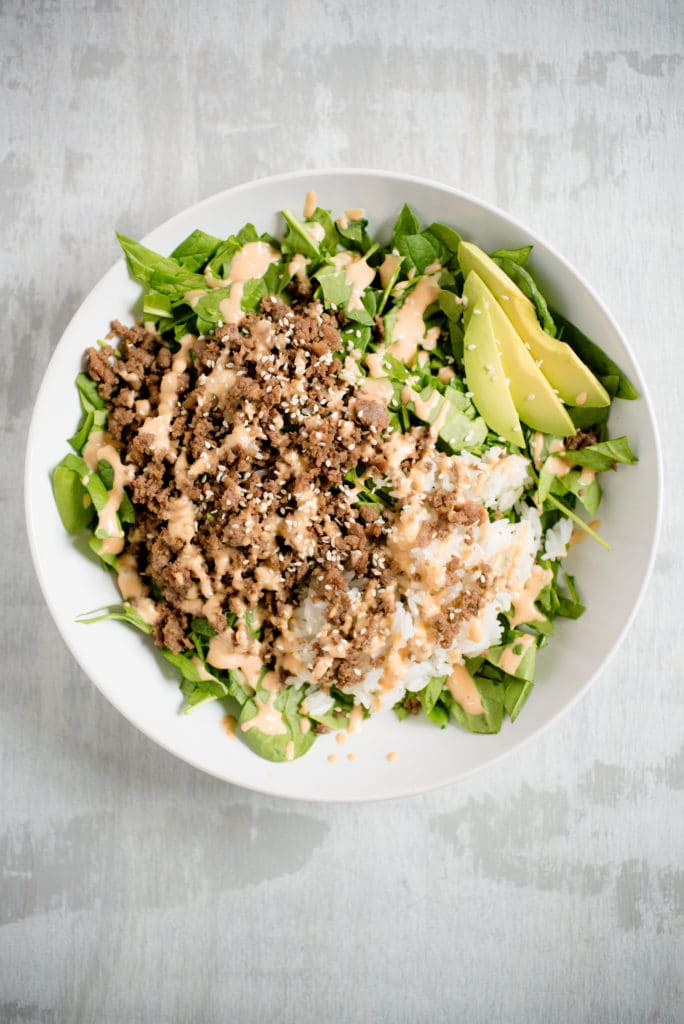 large white bowl with greens, korean beef, white rice, sliced avocado, white sesame seeds, and spicy mayo