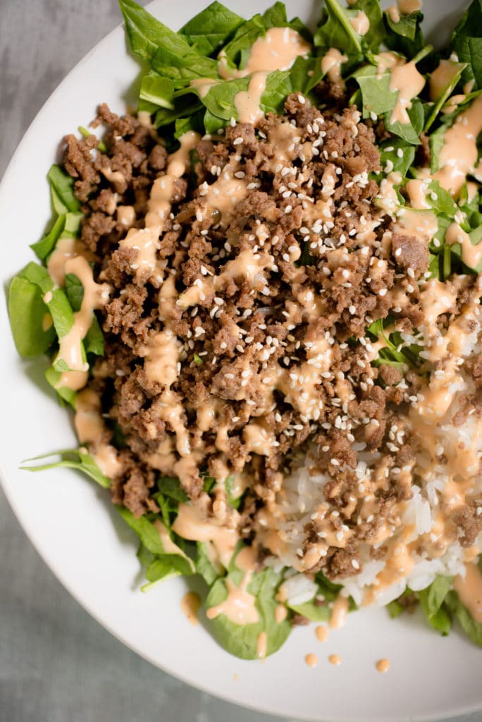lettuce greens in a white bowl, topped with korean ground beef, white rice, spicy mayo and white sesame seeds