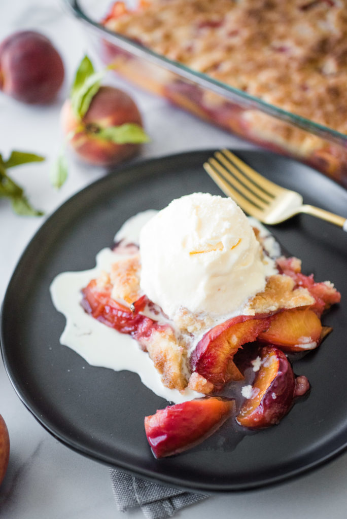 up close shot of peach cobbler on a round, black plate topped with a scoop of vanilla ice cream that is beginning to melt