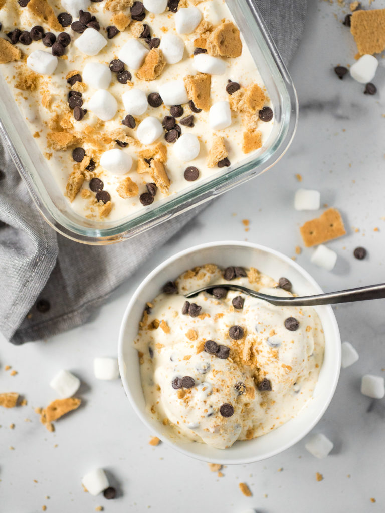 overhead shot of s'mores no churn ice cream topped with marshamallows, chocolate chips, graham crackers. part is in a glass container wile two scoops are in a white bowl