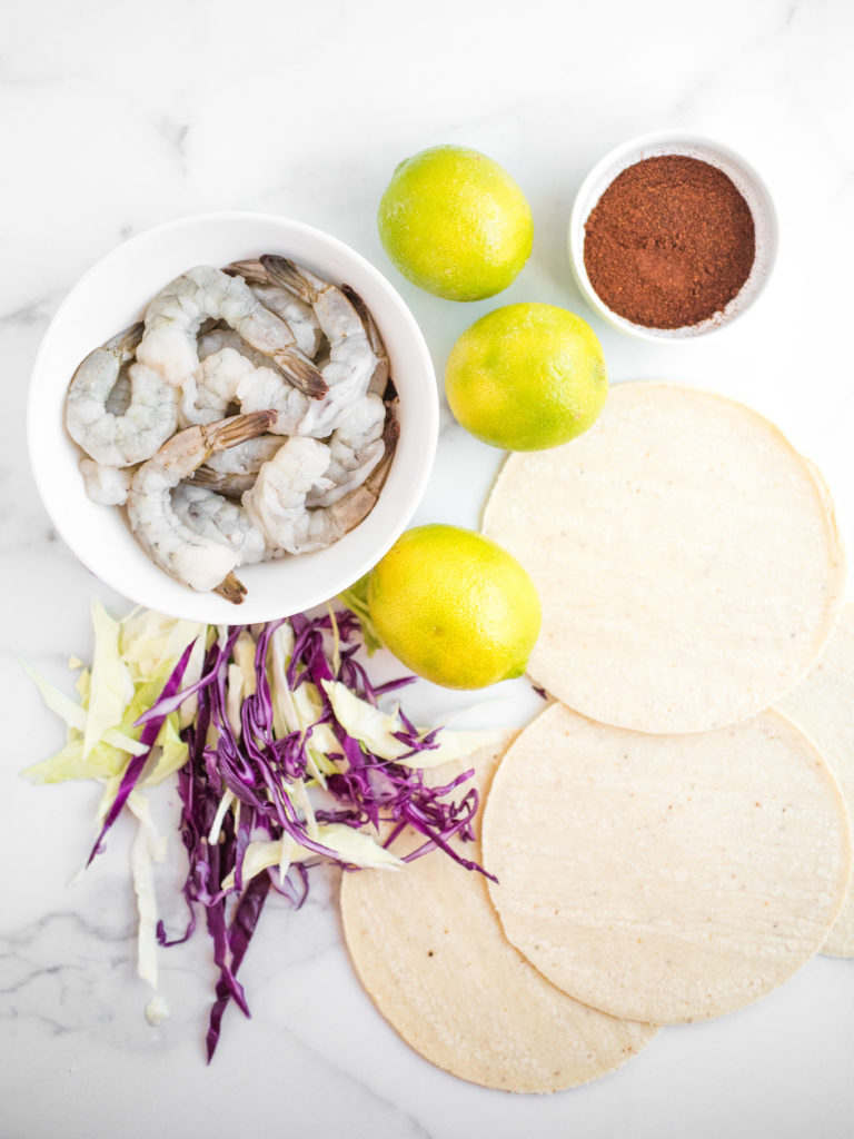 overhead shot of shrimp in a white bowl surrounded by spices, limes, corn tortillas, and shredded cabbage