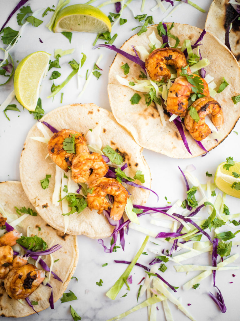 overhead shot of chili lime shrimp tacos in charred white corn tortillas with cabbage slaw surrounded by additional cabbage and limes