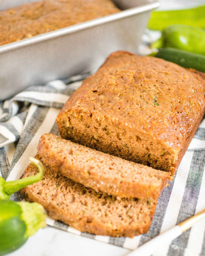 loaf of green chile zucchini bread with two slices cut and laying in front of the rest of the loaf. there is a pan in the background with another loaf.