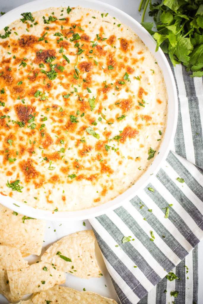 Large, round, shallow white casserole dish with green chile queso blanco dip inside. Topped with cilantro and cheese.