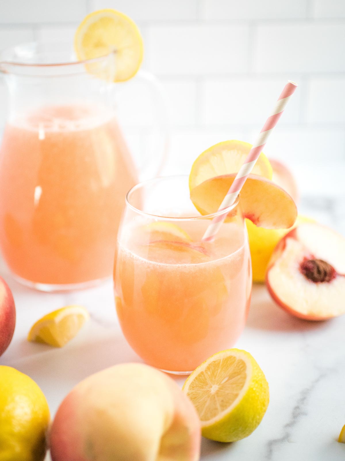 a glass of peach lemonade in front of the pitcher with peaches and lemons on the counter