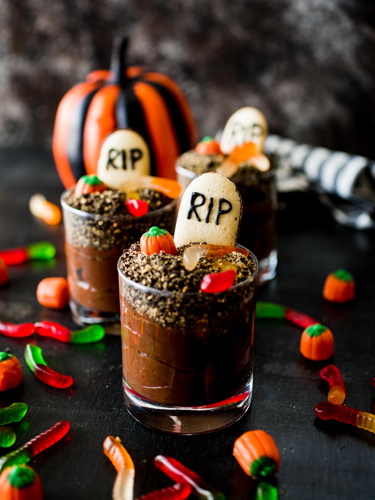 Halloween Graveyard Dirt Cups arranged with an assortment of candies and a small pumpkin, creating a festive spooky scene.