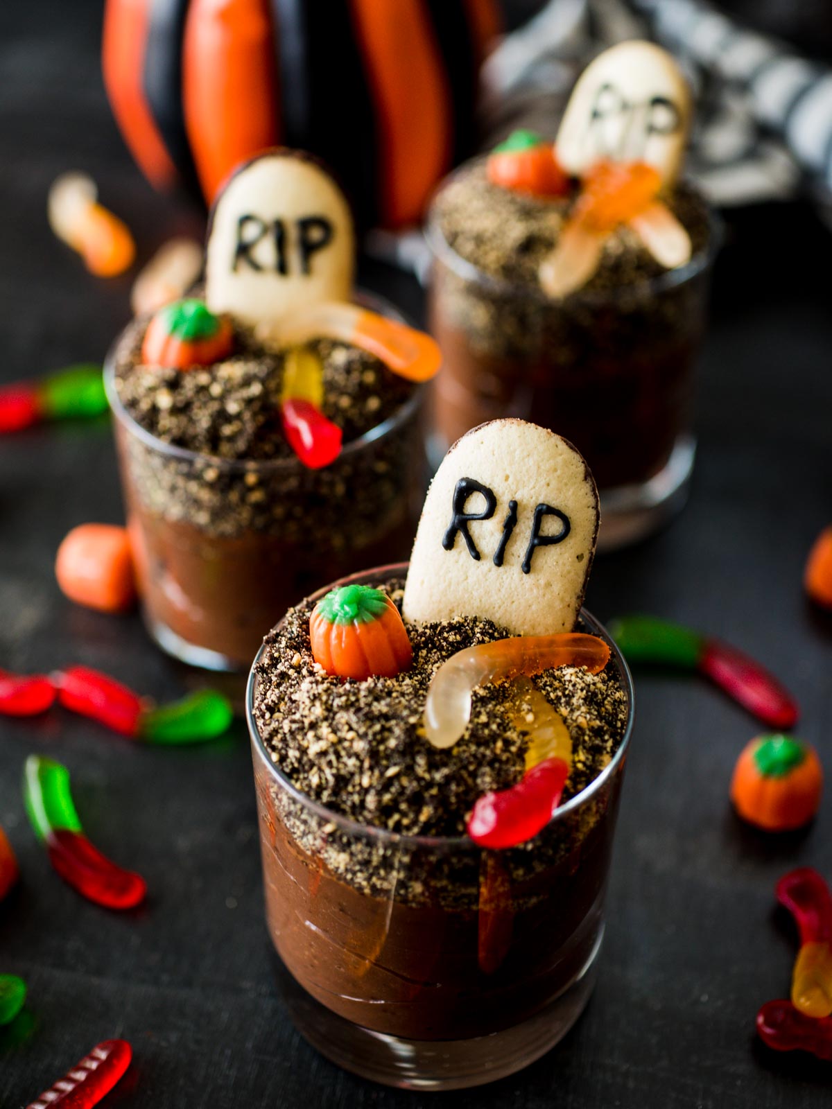 Halloween Graveyard Dirt Cups with gummy worms and mellowcreme pumpkins on top, surrounded by loose candies and cookie crumbs.