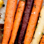 A close-up of roasted carrots in orange, purple, and white varieties, sprinkled with herbs and coarse salt on a baking tray. A label at the top reads, brown sugar roasted carrots, highlighting this brown sugar carrots recipe.