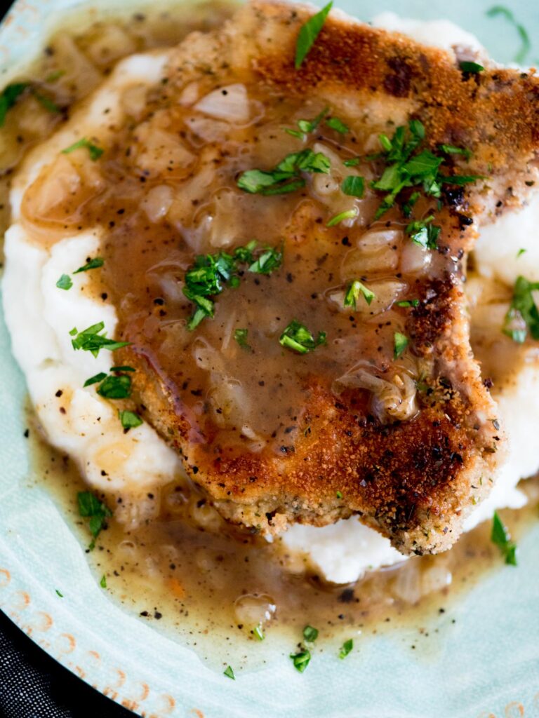 crispy fried pork chops with gravy over a bed of mashed potatoes on a plate