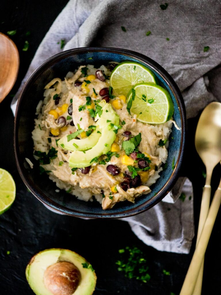 Bowl of salsa verde chicken with lime and avocado slices garnished on top with a spoon resting next to it. 