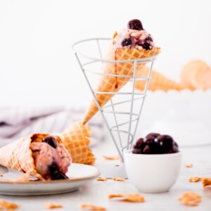 homemade black cherry ice cream in a cone drizzled with cherry juice down the side - a bowl full of cherries on the side