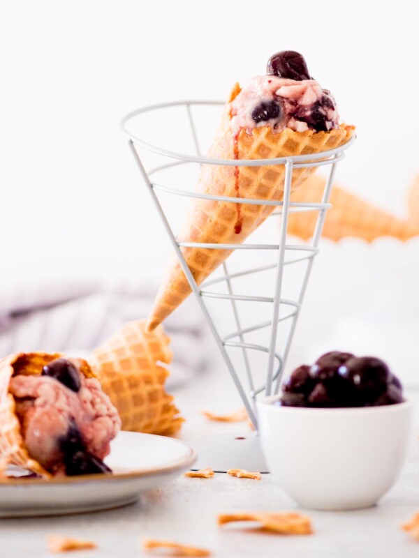 homemade black cherry ice cream in a cone drizzled with cherry juice down the side - a bowl full of cherries on the side