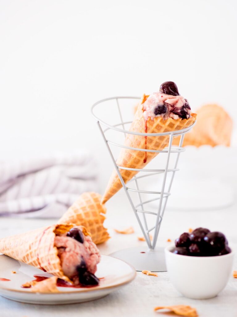 homemade dark cherry ice cream in a cone with a bowl of cherries and another cone on a plate