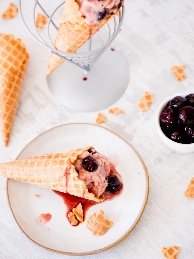 black cherry ice cream in a cone on a plate surrounded by a bowl of cherries and pieces of waffle cone