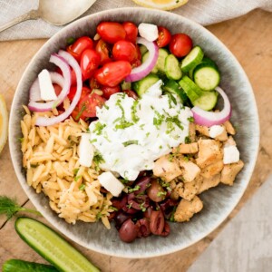 greek chicken bowl topped with tzatziki surrounded by cucumbers and dill