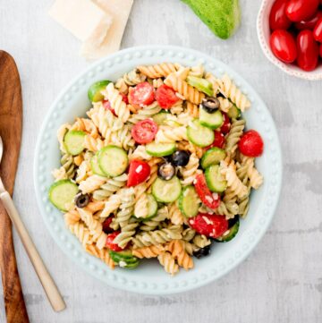 italian pasta salad in a bowl surrounded by parmesan, cucumber, and tomato