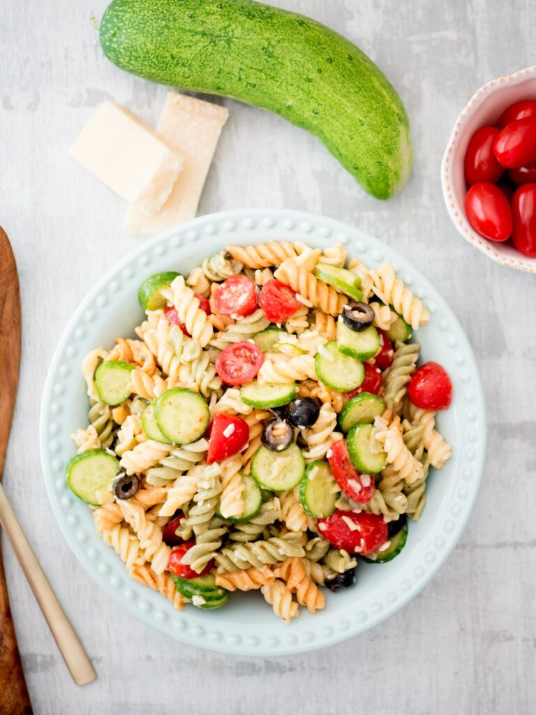 zesty italian pasta salad in a blue bowl surrounded by parmesan cucumber and tomato