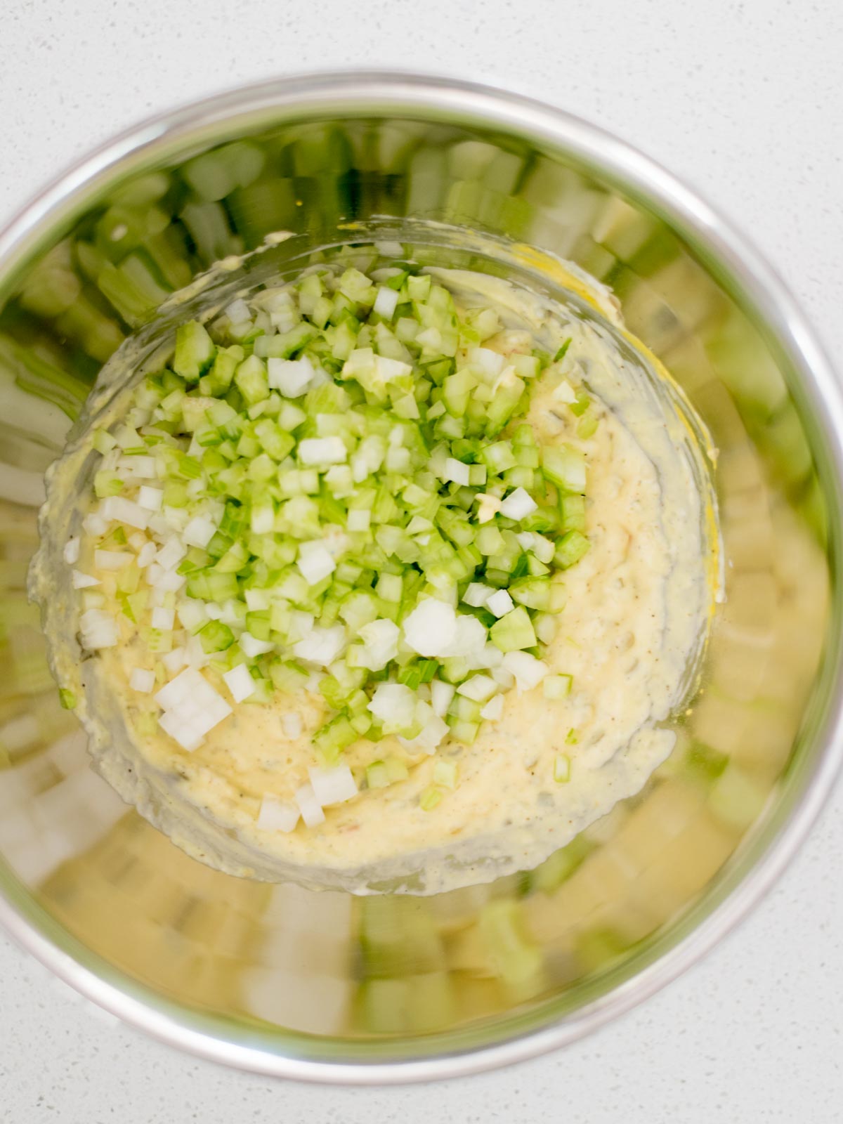 celery and onion added to potato salad dressing in a mixing bowl