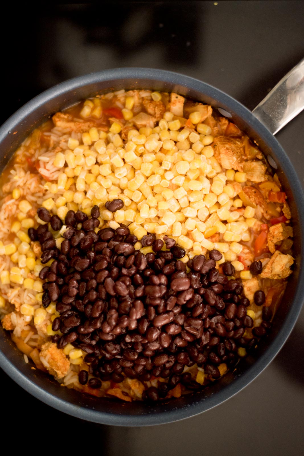 corn and black beans added to the skillet with the cooked chicken, bell peppers, and rice on the stove