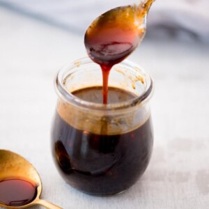 Spoon hovering over a small glass jar of honey sriracha sauce drizzling the dark sauce back in.