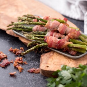Platter of bacon wrapped asapargus with crispy bacon pieces around the edge.