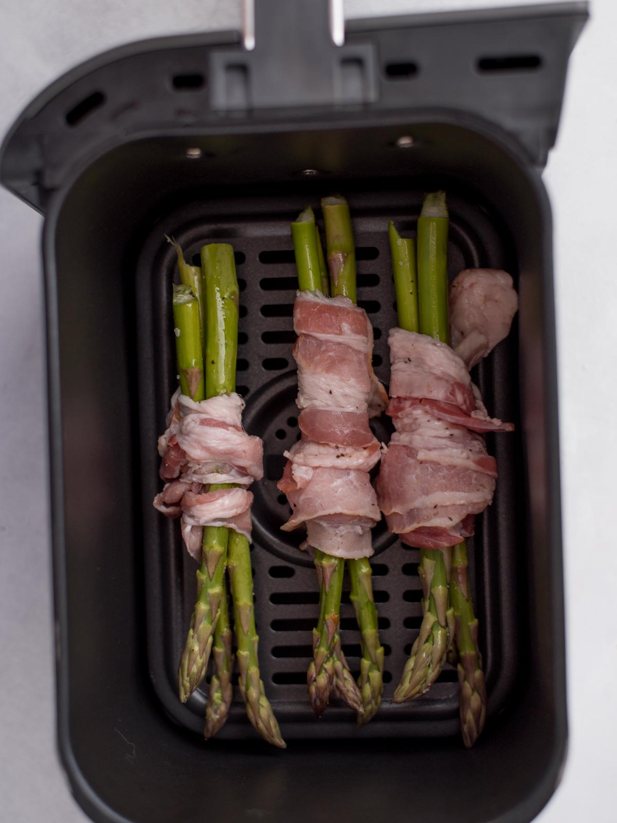 Seasoned asparagus wrapped in bacon and laying in an air fryer basket.