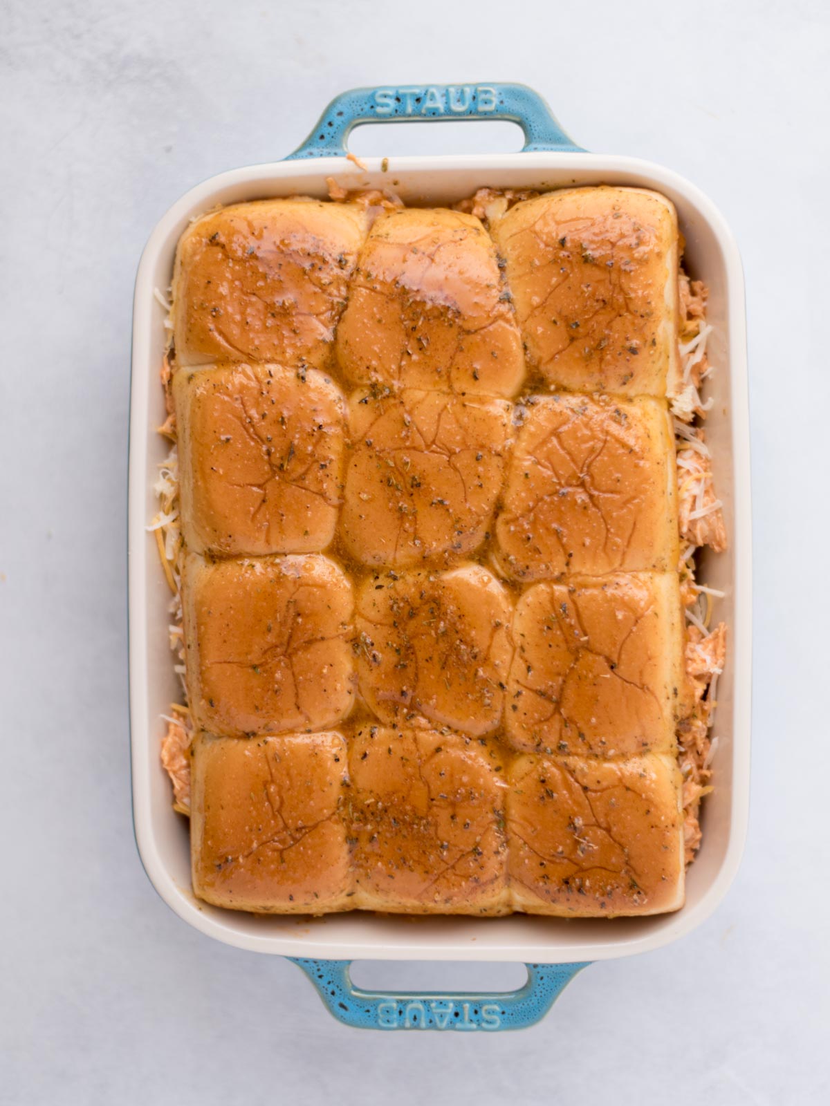 Seasoned melted butter poured over buffalo chicken sliders in a baking dish before baking.