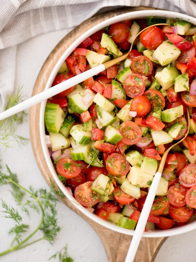 Top view of a large bowl of cucumber tomato salad and salad tongs resting in it.