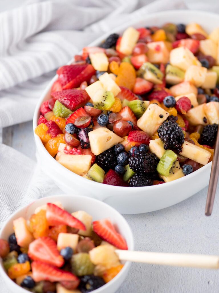 fruit salad in a white bowl with a serving spoon next to a smaller serving of fruit salad in a small white bowl with a fork