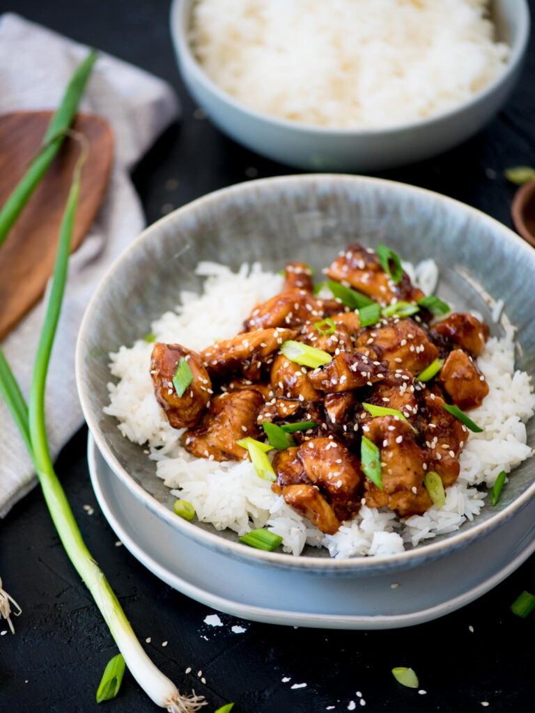 A whole green onion laying next to a bowl of General Tso Chicken and rice.