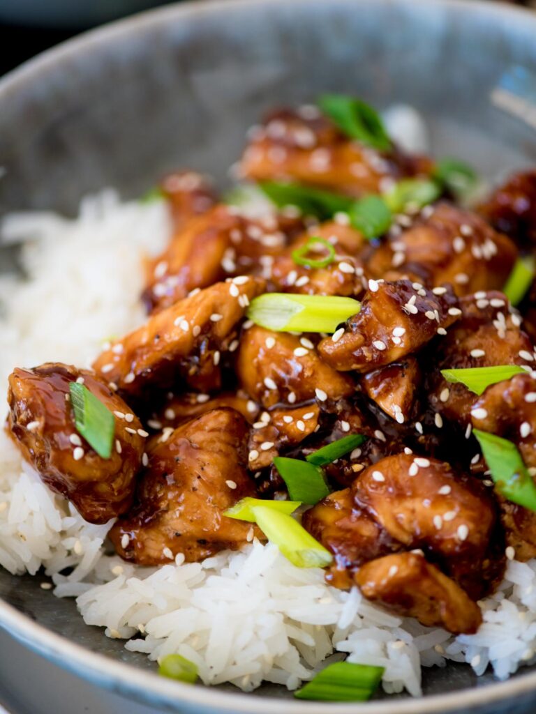 Sliced green onions over General Tso chicken and white rice in a bowl.