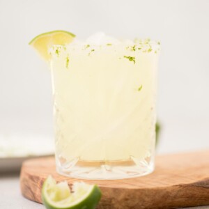 margarita on the rocks in a glass that's lined with salt and lime zest on a wooden board with a lime wedge
