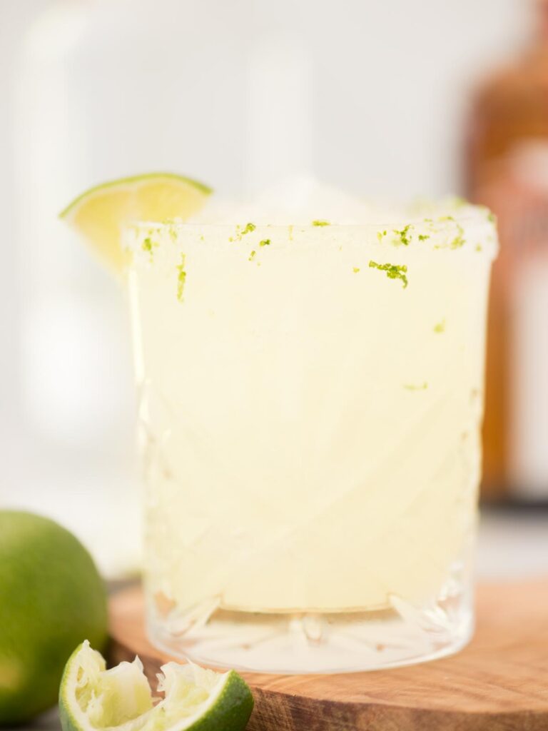 margarita on the rocks in a salt and lime zest rimmed glass and a lime wedge sitting on a wooden board with an extra lime to the side