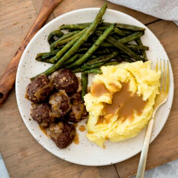Mashed potatoes topped with gravy on a plate with french onion meatballs and green beans.