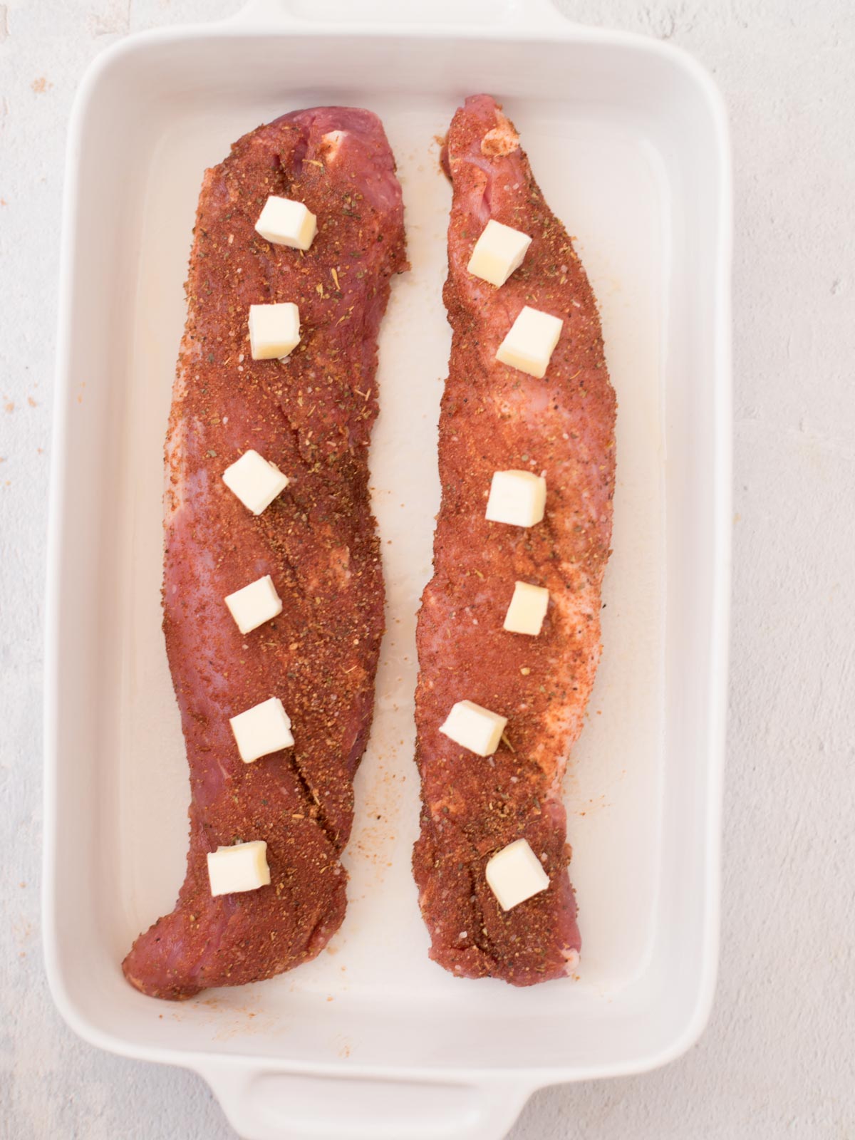raw pork tenderloin in a baking dish seasoned and topped with cubes of butter