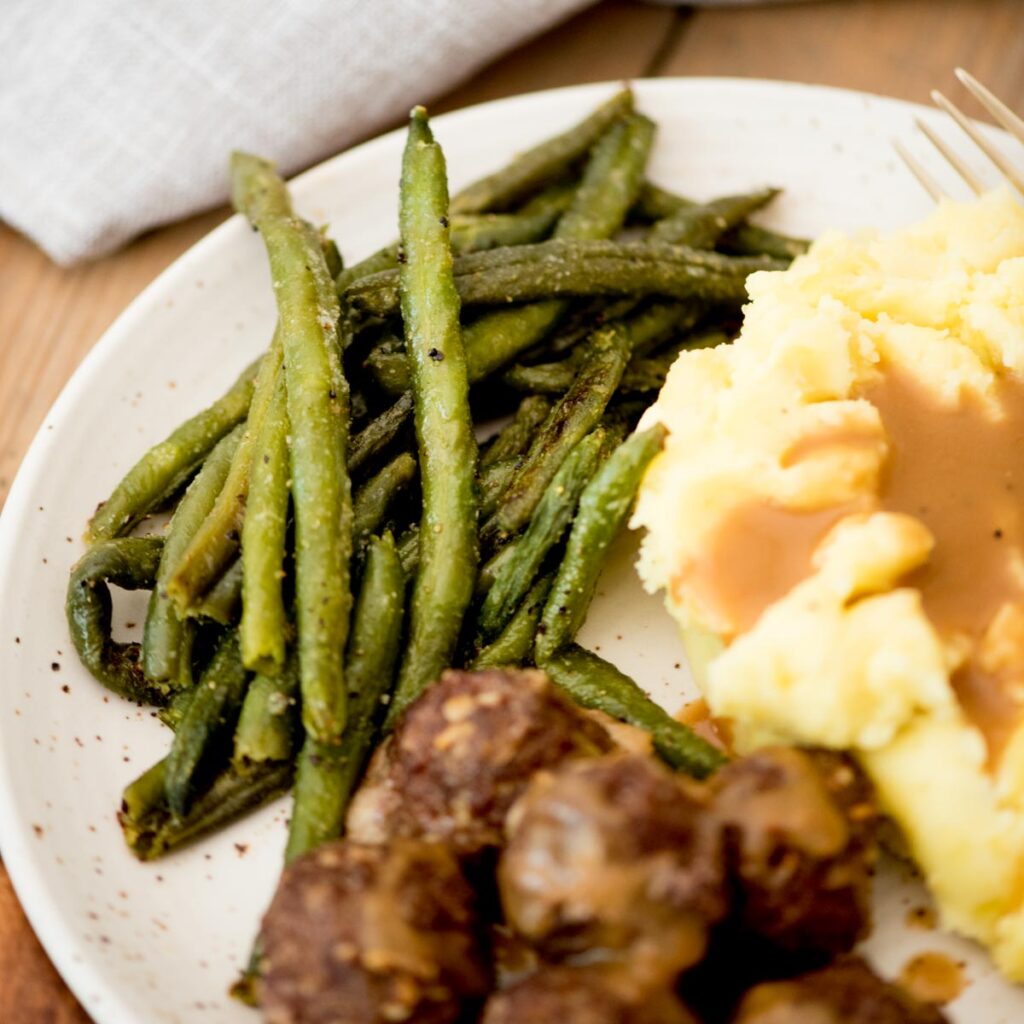 A side of roasted frozen green beans on a plate with meatballs and mashed potatoes.