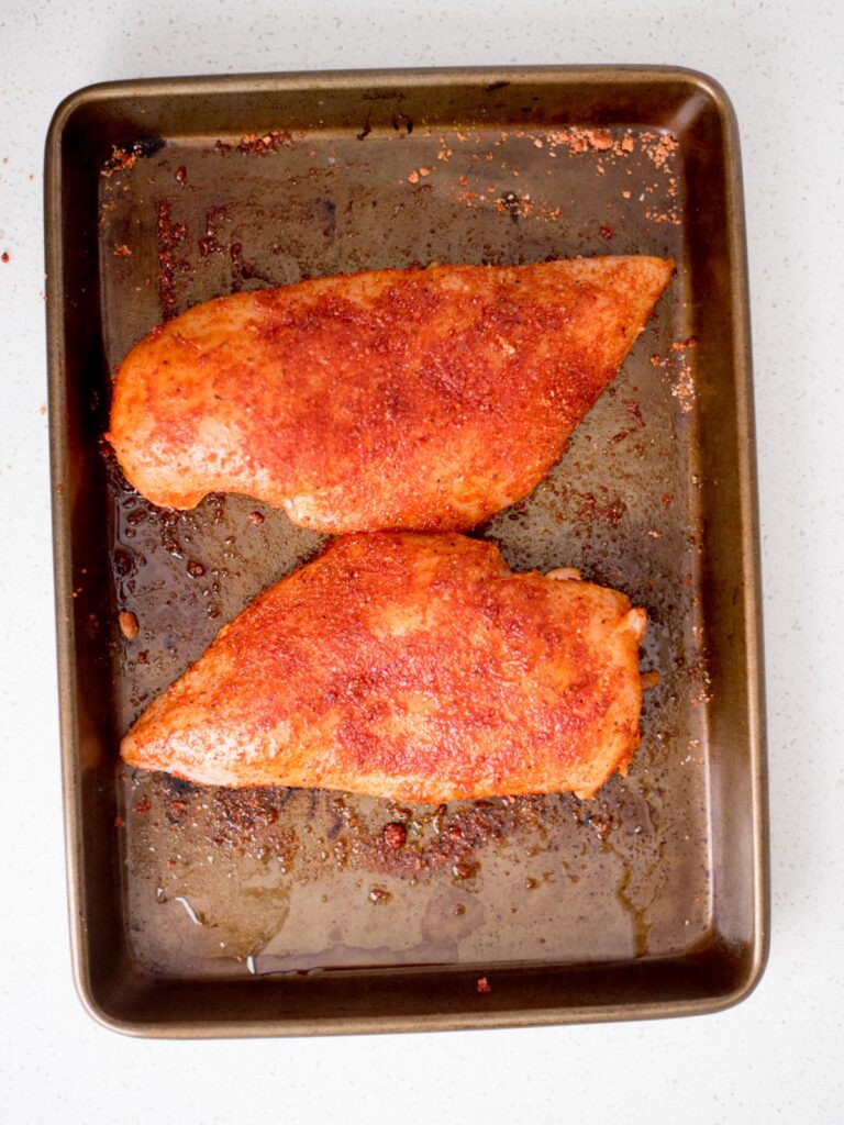 Two uncooked seasoned chicken breasts on a baking sheet.