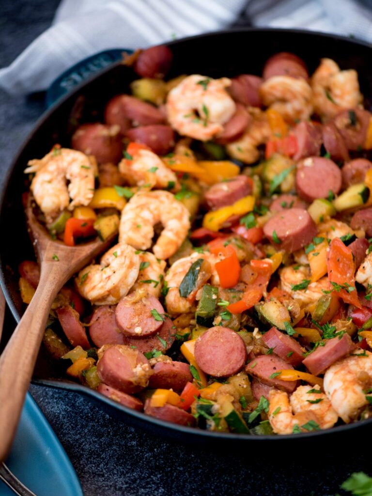 Wooden spoon holding a scoop of cajun shrimp and sausage with vegetables in a cast iron pan.