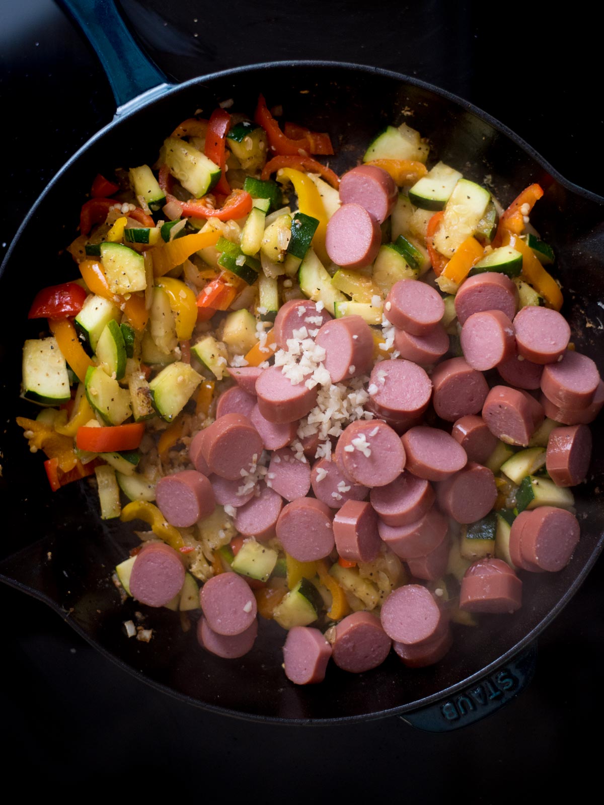Veggies and sausage added to a skillet with diced garlic.