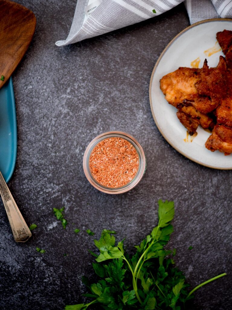 Small glass jar of chicken seasoning next to a plate of cooked chicken wings and fresh herbs