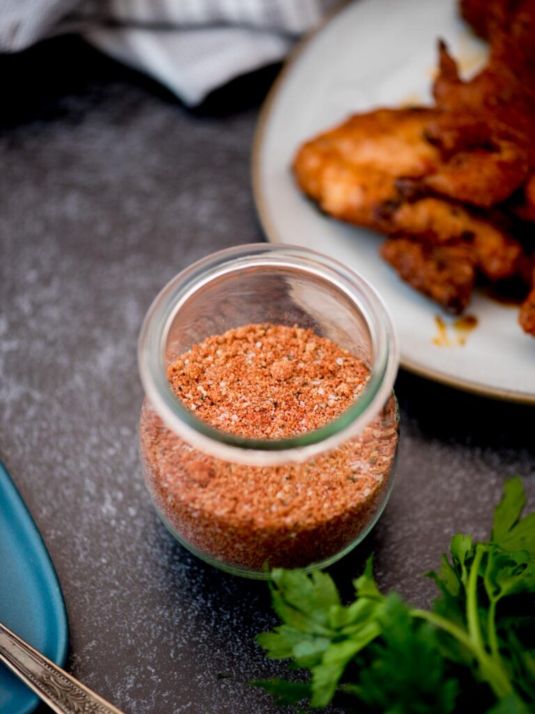 Small glass jar of chicken seasoning next to a plate of chicken wings.