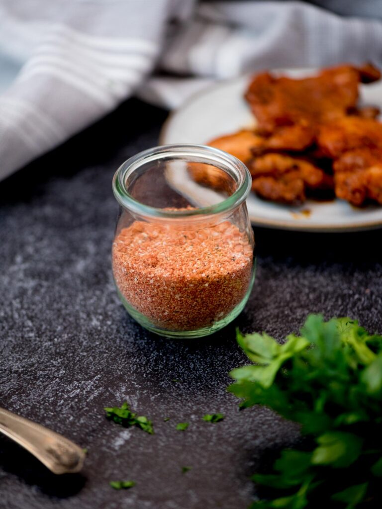 Fresh herbs in front of a glass of chicken seasoning and a plate of cooked chicken wings