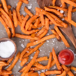 Sea salt sprinkled over sweet potato fries on parchment paper with a small dish of ketchup and sea salt by them.