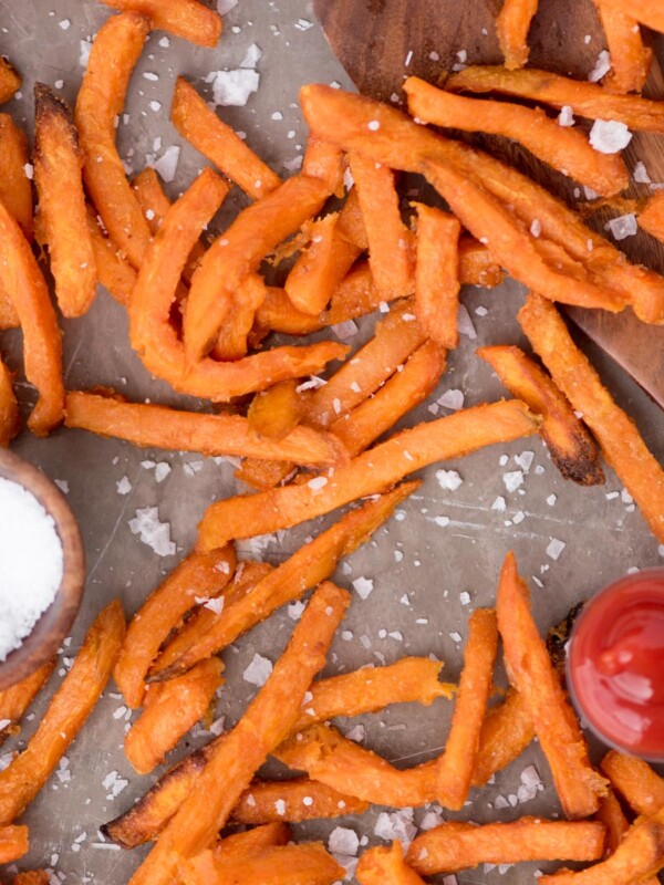 Sea salt sprinkled over sweet potato fries on parchment paper with a small dish of ketchup and sea salt by them.