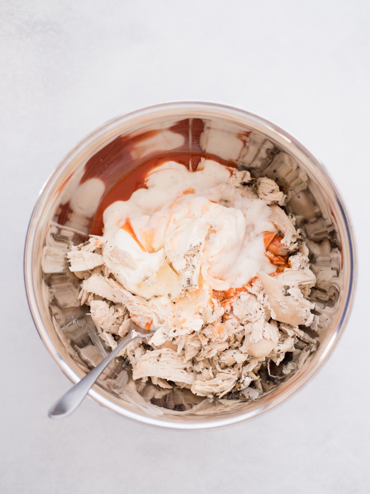 Shredded chicken and ingredients for buffalo chicken added to a mixing bowl with a spoon.