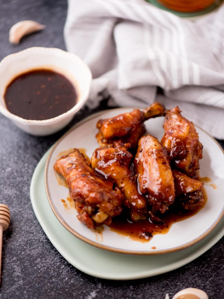 honey garlic chicken wings stacked on a white plate with a bowl of extra sauce and garlic cloves