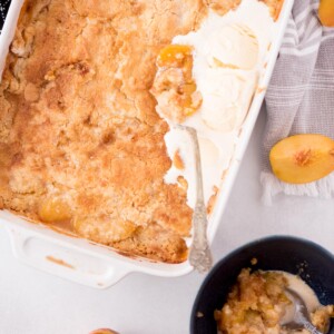 Peach cobbler with a scoop of ice cream melting in the baking dish and a small bowl served of the dessert next to it.